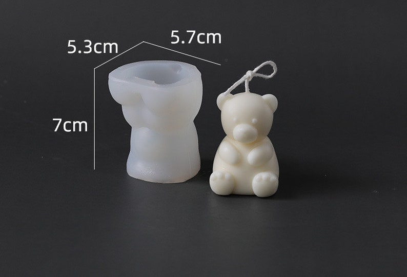 May include: A white bear-shaped candle and a silicone mold. The candle is 5.7 cm tall. The mold is 7 cm tall and 5.3 cm wide. The candle has a string attached. The items are on a dark surface.