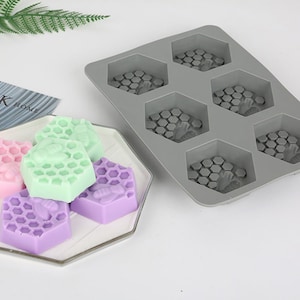 May include: A gray silicone mold with six hexagonal cavities, each featuring a honeycomb pattern with a bee design.  Four hexagonal soaps are shown in the foreground, two purple, one green, and one pink, each with a honeycomb pattern and a bee design.