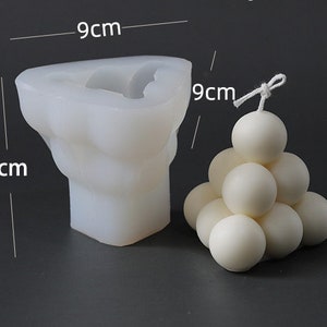 May include: A white silicone candle mould, 8cm x 9cm, next to a finished white sphere candle. The mould is in the shape of a torso. The sphere candle is made of multiple spheres stacked in a pyramid shape. Candle making supplies.