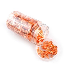 May include: A clear plastic jar filled with copper leaf flakes. The jar is tipped over and some of the flakes are spilling out.