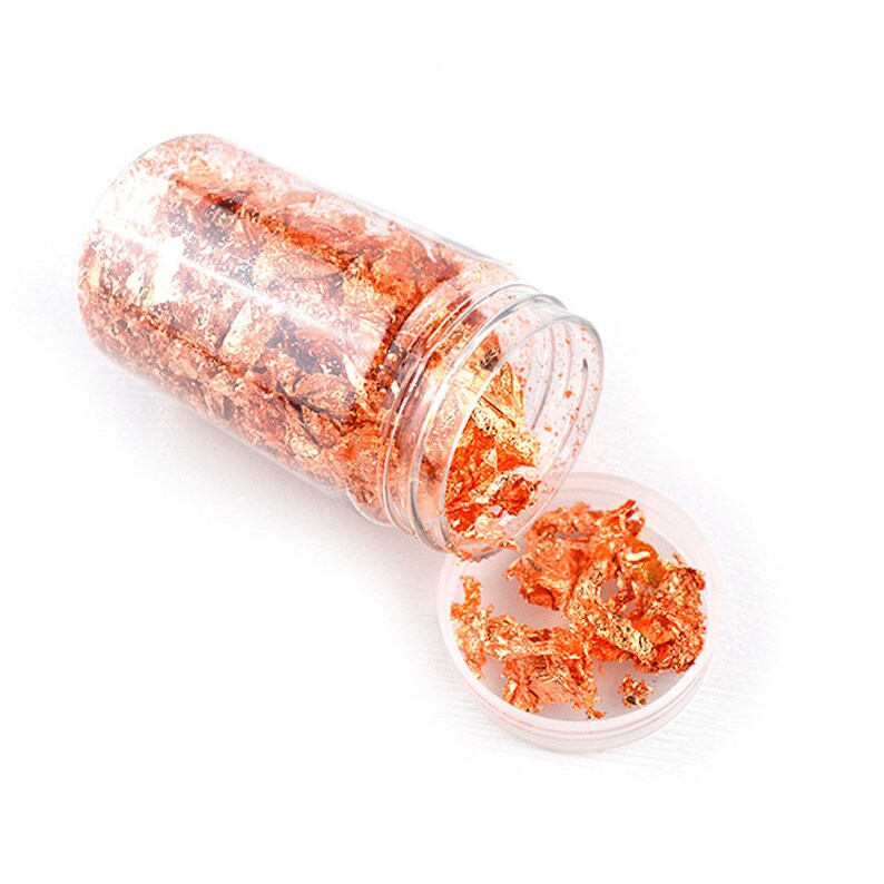 May include: A clear plastic jar filled with copper leaf flakes. The jar is tipped over and some of the flakes are spilling out.