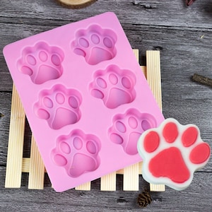 Peut inclure: Moule en silicone rose avec six formes de pattes d'animaux. Une empreinte de patte rouge est posée sur une surface en bois à côté du moule.