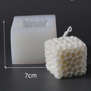 May include: A white, cube-shaped candle with a honeycomb pattern, next to a silicone mold. The candle measures approximately 7 cm wide and 6 cm tall. A white wick is visible at the top of the candle. The mold is 7 cm wide and 6 cm tall.
