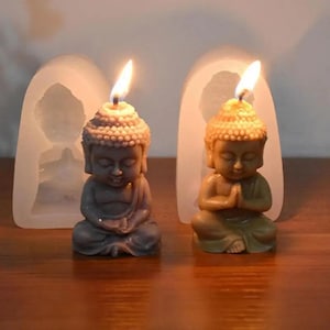 May include: Two small, detailed Buddha candle molds made of silicone. The molds are white and have a smooth surface. The Buddha candles are brown and gray and have a flame on top.