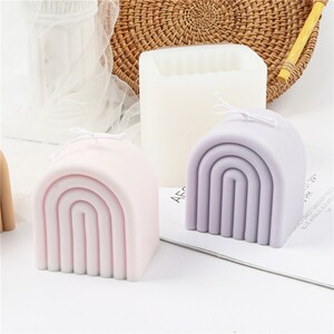 May include: Two rainbow-shaped silicone moulds for making candles. The moulds are in pink and purple and are sitting on a white surface.