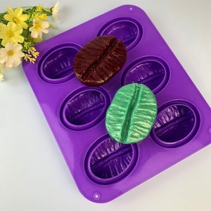 Op de afbeelding: Paarse siliconen mal met acht holtes gevormd als koffiebonen. Twee koffieboonvormige zepen bevinden zich in de mal, een is donkerbruin en de andere is lichtgroen.