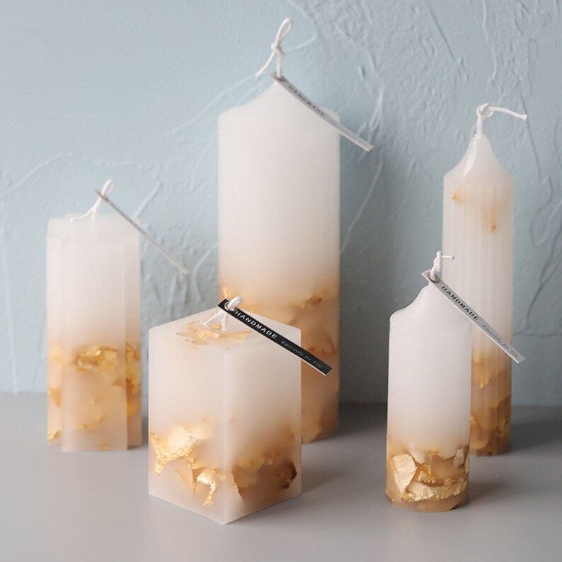 May include: Five white pillar candles with gold leaf accents. The candles are of varying heights and are arranged on a light blue surface. Each candle has a small black tag with the word "HANDMADE" printed on it.