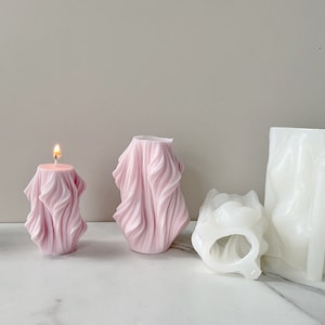 Puede incluir: Tres velas decorativas, dos son rosas y una es blanca. Las velas rosas tienen un diseño ondulado y texturizado. La vela blanca tiene una forma más abstracta y orgánica.