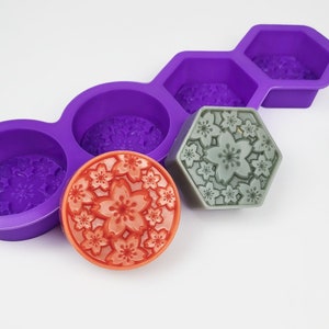 May include: A purple silicone mould with four round and one hexagonal cavities, each with a floral design. Two finished soap bars are shown, one round and orange, the other hexagonal and grey.