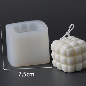 May include: A white, cube-shaped candle next to a silicone mould. The candle has a textured, bubble-like design. The mould is 6 cm tall and 7.5 cm wide. The candle has a white string attached. The background is a solid grey.
