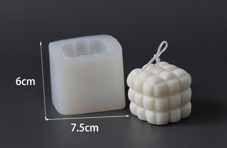 May include: A white, cube-shaped candle next to a silicone mould. The candle has a textured, bubble-like design. The mould is 6 cm tall and 7.5 cm wide. The candle has a white string attached. The background is a solid grey.