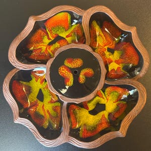 May include: A ceramic serving dish with a flower-like design. The dish features five sections with a central circular area. The interior of each section has a vibrant pattern of yellow, orange, and red. The outer rim is a brown, wood-like texture.