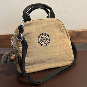 May include: Tan nylon handbag with a black handle and shoulder strap. The bag features a round logo patch and a small monkey keychain. The bag is designed with a zip closure and is suitable for everyday use.