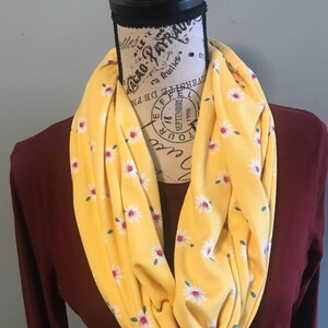 May include: A yellow infinity scarf with a white daisy print. The daisies have red centers and green leaves.
