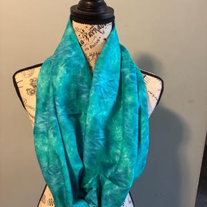May include: A teal and turquoise infinity scarf displayed on a mannequin. The scarf has a tie-dye pattern with shades of blue and green. The fabric is draped in soft folds, showcasing the texture and design.
