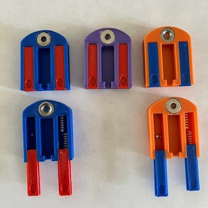 May include: Five U-shaped plastic clips with rounded tops and metal eyelets. Three clips are closed with red or blue inserts, while two clips have extended spring-loaded arms. The colors include blue, purple, and orange plastic components.