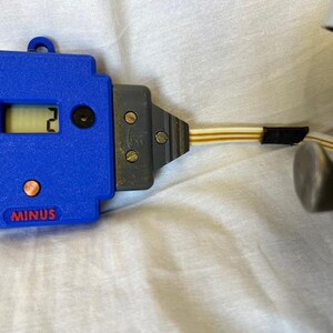 May include: A blue digital device with a display showing the number 2. The device has a gray connector and a cable with a black tape. The word "MINUS" is printed on the device.