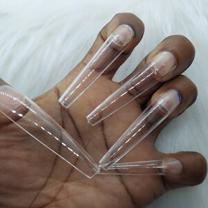 May include: A set of five clear plastic nail tips, designed for nail extensions. The tips are long and narrow, with a rounded tip. They are held in a hand.