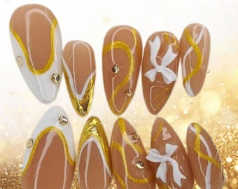 Uñas postizas blancas Coquette con lazos dorados, ideales para novias y graduaciones.