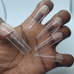 May include: Clear, rectangular nail tips in various sizes are displayed on a hand. The tips are transparent, showing the natural nail underneath. Each tip has a number indicating its size, and is ready for application.
