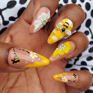 Nude yellow mustard colored nails with white flowers drawn on thumb and pinky nail. middle finger has a 3d bee charm on yellow checkered nail with rhinestones. other two nails have 3d yellow and white acrylic flowers.