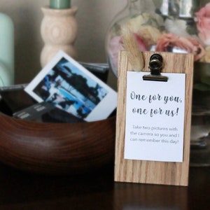 May include: A wooden sign with a clip holding a white card that says "One for you, one for us! Take two pictures with the camera so you and I can remember this day!"