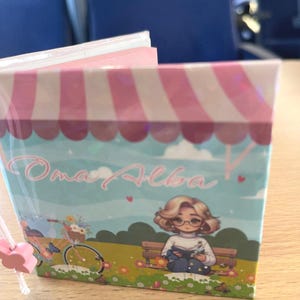 Puede incluir: Un toldo a rayas rosa y blanco con el texto "Oma Alba" en cursiva. Una ilustración de dibujos animados de una mujer con cabello rubio y gafas sentada en un banco leyendo un libro. Una bicicleta con una cesta de flores está en el fondo.
