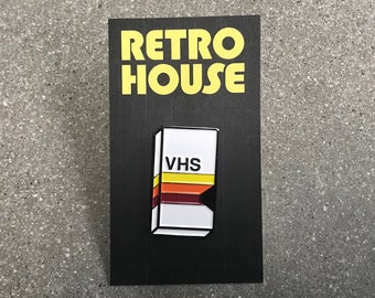 May include: A black and white enamel pin featuring a VHS tape with a yellow, orange, and red stripe design. The pin says "VHS" in black text. The pin is on a black card with the text "RETRO HOUSE" in yellow text.
