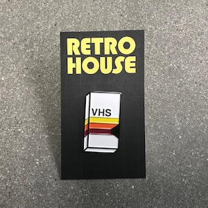 Könnte beinhalten: Eine schwarz-weiße Emaille-Anstecknadel mit einem VHS-Band mit einem gelben, orangefarbenen und roten Streifenmuster. Die Nadel trägt den Schriftzug "VHS" in schwarzer Schrift. Die Nadel befindet sich auf einer schwarzen Karte mit dem Schriftzug "RETRO HOUSE" in gelber Schrift.