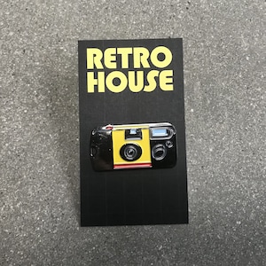 May include: A black and yellow enamel pin featuring a vintage disposable camera with a red button. The pin says "Retro House" in white letters on a black background.