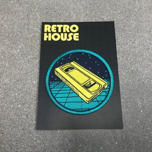 May include: A black patch with the text "RETRO HOUSE" in yellow. The patch features a yellow VHS tape design inside a teal and navy blue circle with a grid pattern, evoking a retro aesthetic.
