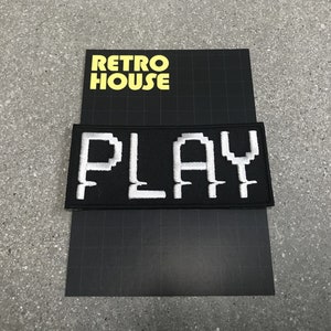 May include: Black and white embroidered patch with the word "PLAY" in pixelated font. The patch is on a black background with a white grid pattern. The text "RETRO HOUSE" is printed in yellow on the background.