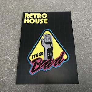 May include: A yellow triangle warning sign with a black and gray robotic hand raised in the air. The text "It's So Bad" is written in pink, black, and blue letters.