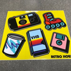 May include: A yellow sticker featuring retro graphics of a camera, roller skate, a 90s style cup, VHS tape, and a floppy disk. The text "RETRO HOUSE" is at the bottom.