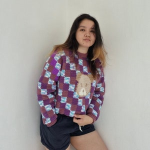May include: A purple, blue and white checked crochet sweater with a brown bear appliqu&eacute;. The sweater has a crew neck and long sleeves.
