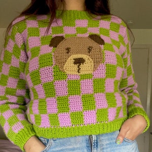 May include: A green and purple checkered crochet sweater with a brown bear applique on the front.