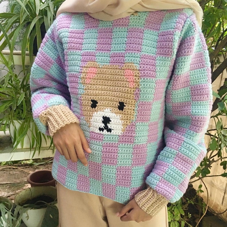 May include: A light purple and blue crocheted jumper with a brown bear face design. The jumper has a checkered pattern.