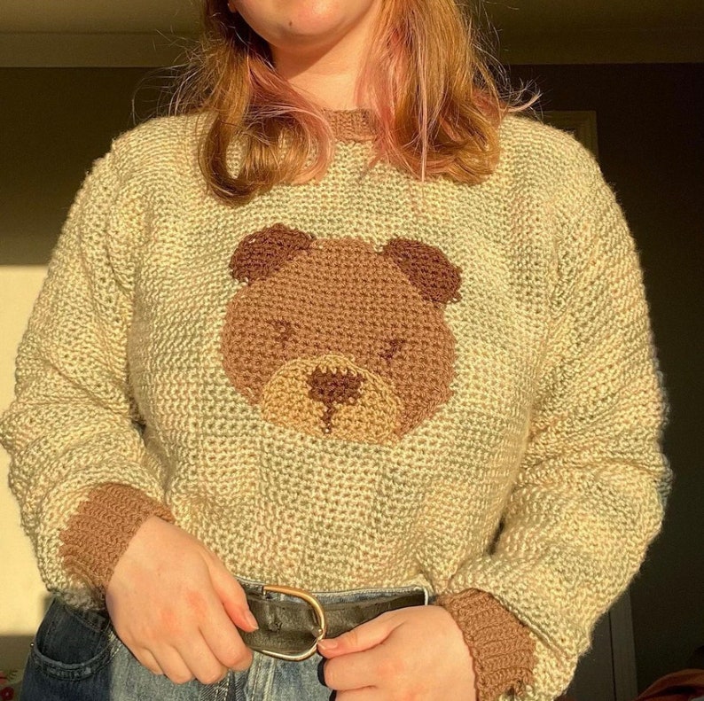 May include: A light green knitted jumper with a brown bear face embroidered on the front. The jumper has brown ribbed cuffs and a brown ribbed hem.