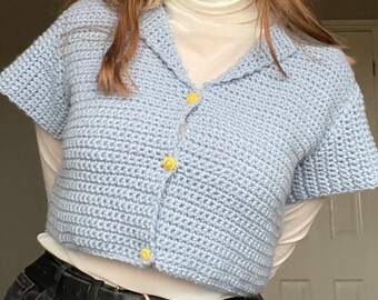 May include: A light blue crocheted cardigan with short sleeves and yellow buttons. The cardigan is cropped and has a collar.
