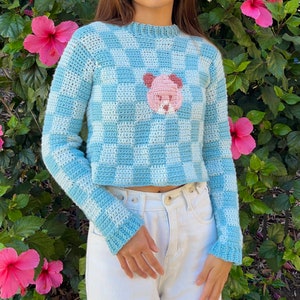 May include: A blue and white gingham cropped sweater with a pink bear applique. The sweater has long sleeves and a crew neck.