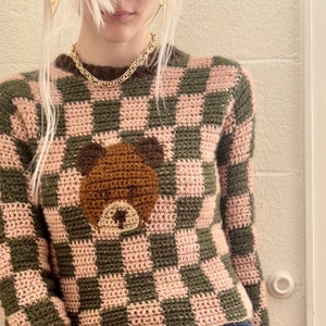May include: A pink and green checked crochet sweater with a brown crocheted bear applique on the front.