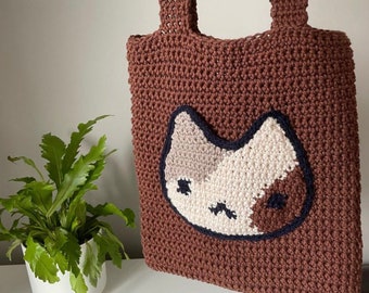 May include: A brown crocheted tote bag featuring a white and brown cat face design. The cat face has black eyes and a black nose.