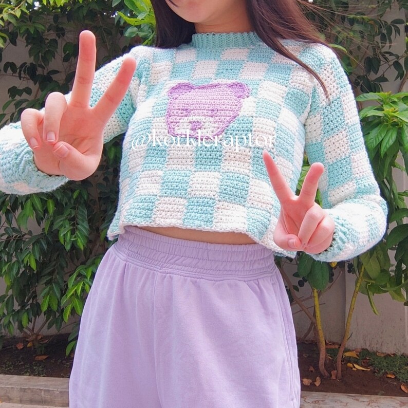 May include: A light blue and white gingham cropped sweater with a purple bear applique. The sweater has long sleeves and a round neckline.