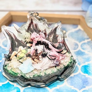 Könnte beinhalten: Ein transparenter Harz-Anhänger mit einer farbenfrohen Anime-Illustration von drei Figuren in einer Waldlandschaft. Der Anhänger liegt auf einem Tablett mit blau-weißem Muster.