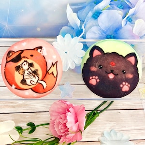 May include: Two round, colorful, decorated cookies. One cookie features a red fox with a black eye patch and a white rabbit. The other cookie features a black cat with a red nose and white paws.