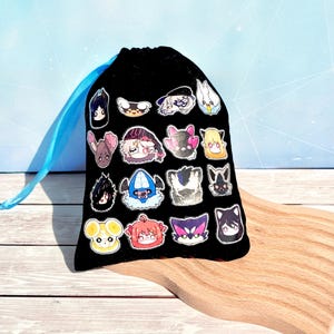 Puede incluir: Una bolsa negra con cordón adornada con pegatinas de personajes de dibujos animados de colores. La bolsa tiene un cordón azul claro y está sobre una superficie de madera. Las pegatinas presentan varias caras de estilo anime.