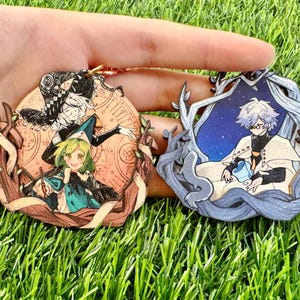 Może przedstawiać: Dwa okrągłe drewniane breloki z ilustracjami w stylu anime. Lewy brelok przedstawia dwie postacie w kapeluszach na tle brzoskwiniowej tarczy zegara. Prawy brelok ukazuje postać w białej pelerynie w niebieskiej, gwiaździstej ramce z drzew.