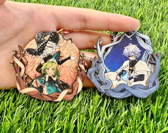 May include: Two circular wooden keychains featuring anime-style illustrations. The left charm shows two characters in hats against a peach-colored clock face. The right charm depicts a character in a white cloak within a blue, starry, tree-framed border.