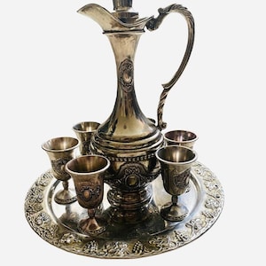 May include: A silver-plated serving set with a pitcher and six cups on a decorative tray. The tray features a grapevine design.