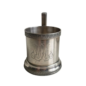 May include: A silver-colored metal cup with a handle. The cup has an engraved monogram on the front and decorative trim around the top and base. The handle is rectangular and attached to the side of the cup.